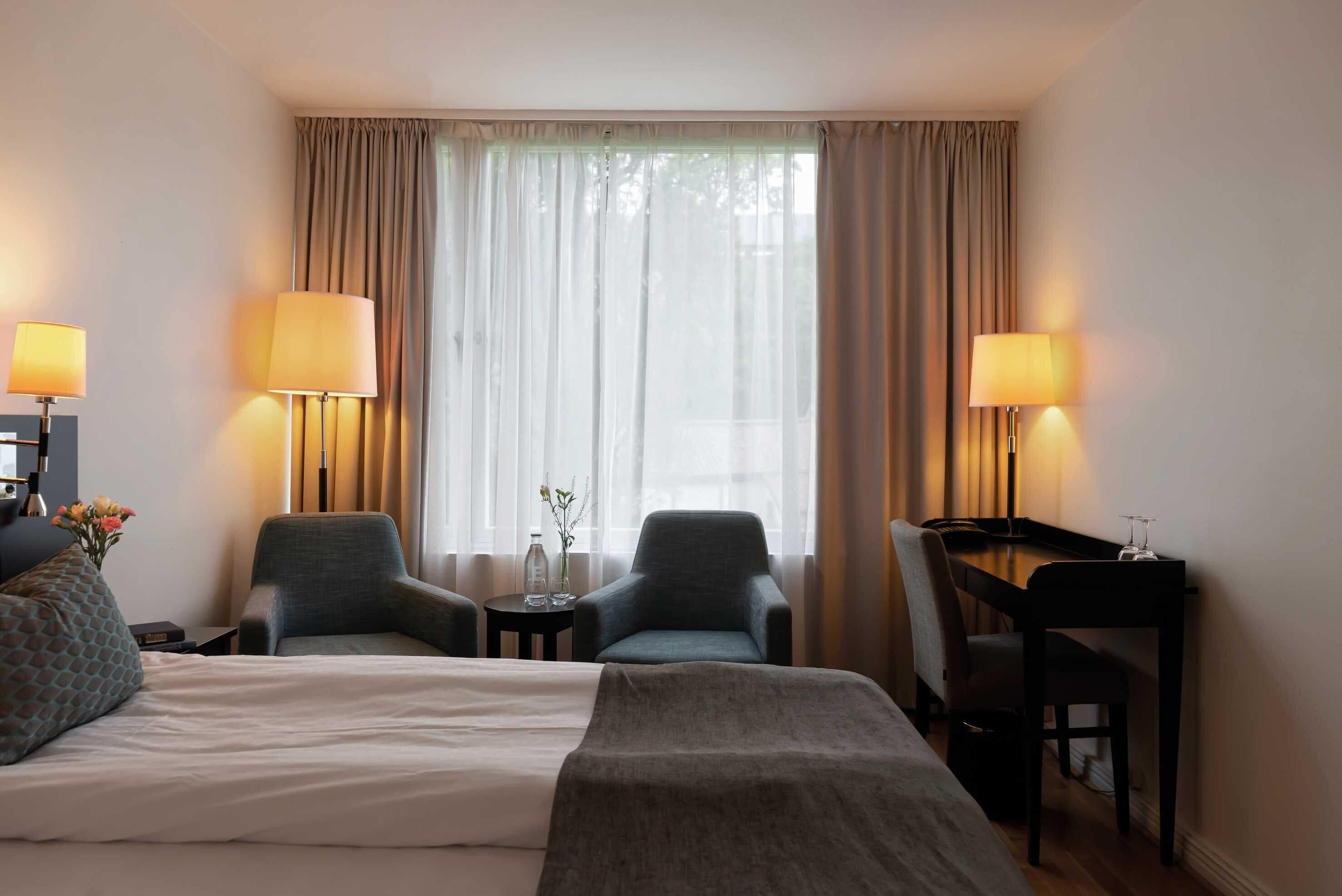 A hotel room with a bed, two armchairs, a desk, and lamps.