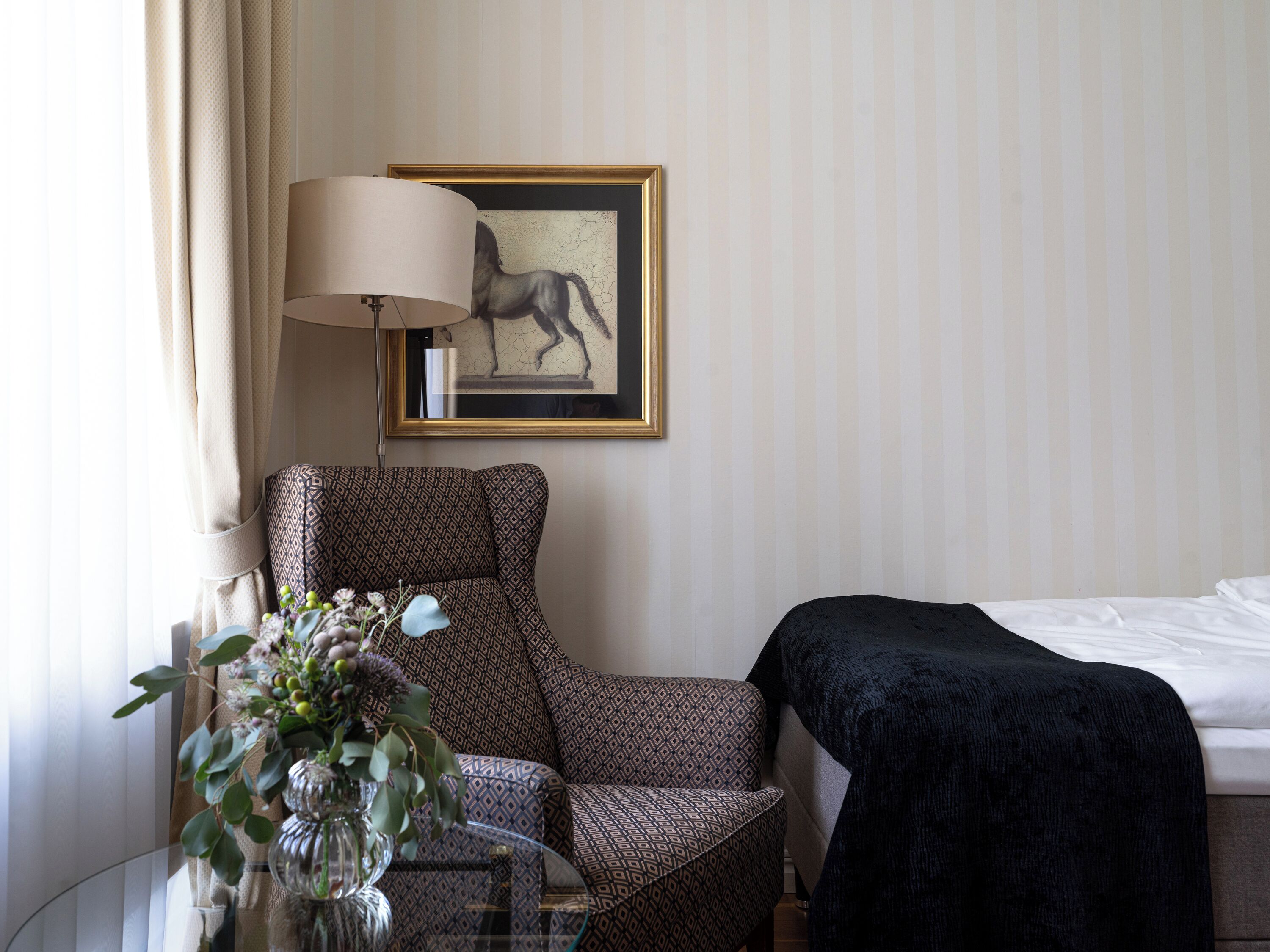 Una habitación de hotel con un sillón estampado, una mesita de noche con un florero y una lámpara, además de un cuadro en la pared.