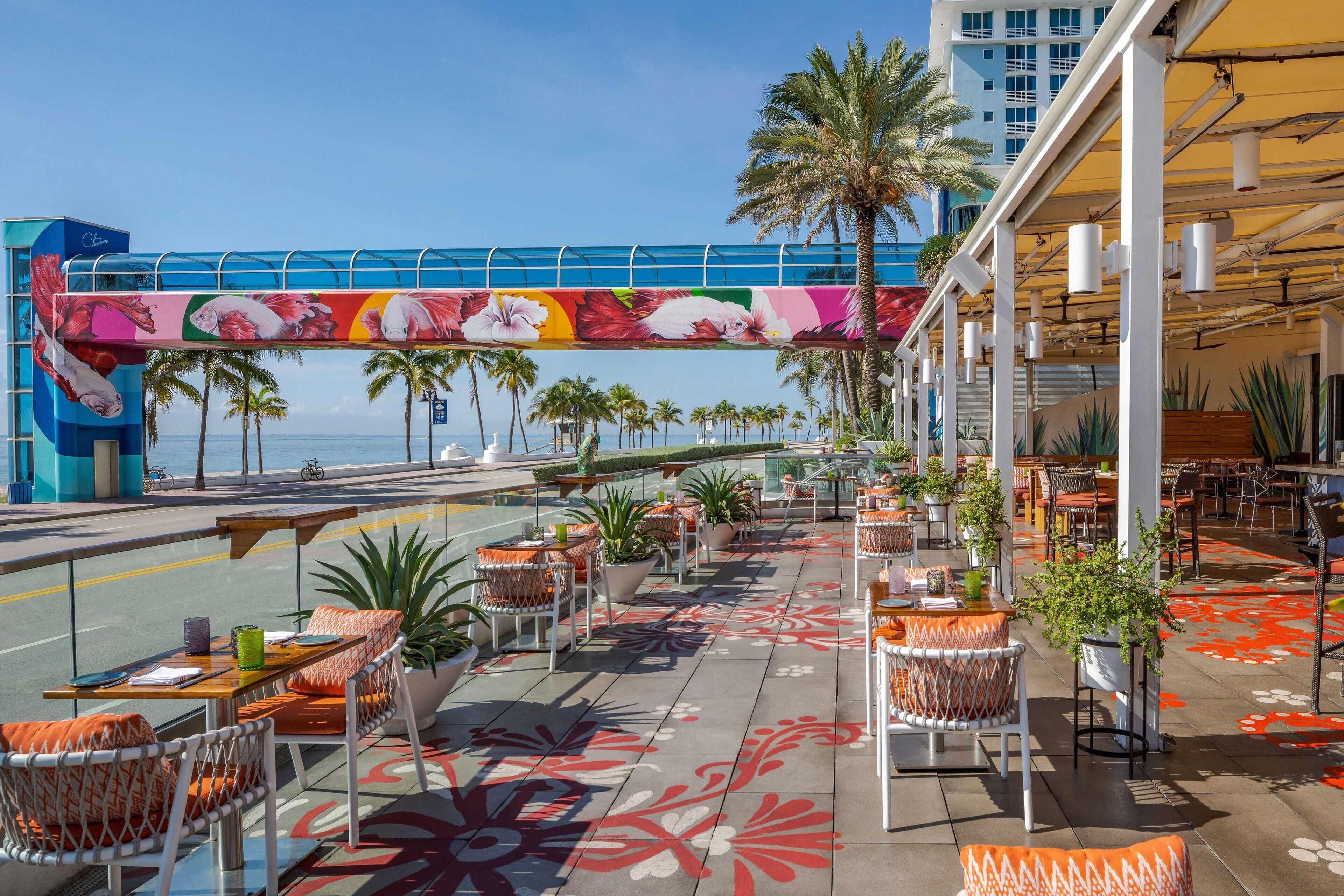 An outdoor seating area with tables and chairs, palm trees, and a view of the ocean.