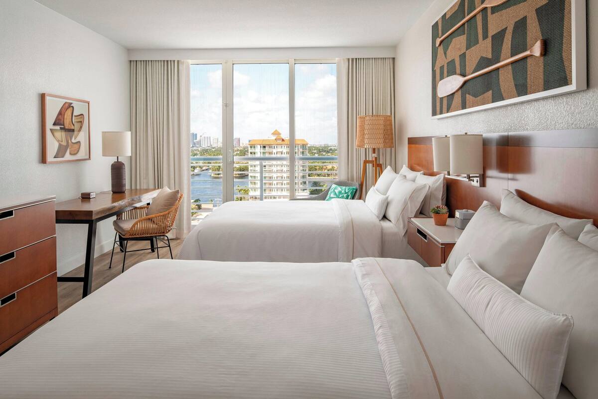 Une chambre d’hôtel avec deux lits, un bureau, une chaise, une grande fenêtre donnant sur la ville et une œuvre d’art accrochée au mur.