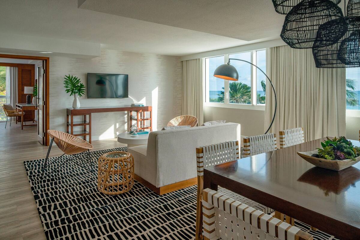 Un salon moderne comprenant un canapé, une table à manger et des chaises, un téléviseur fixé au mur et une grande fenêtre avec des rideaux.