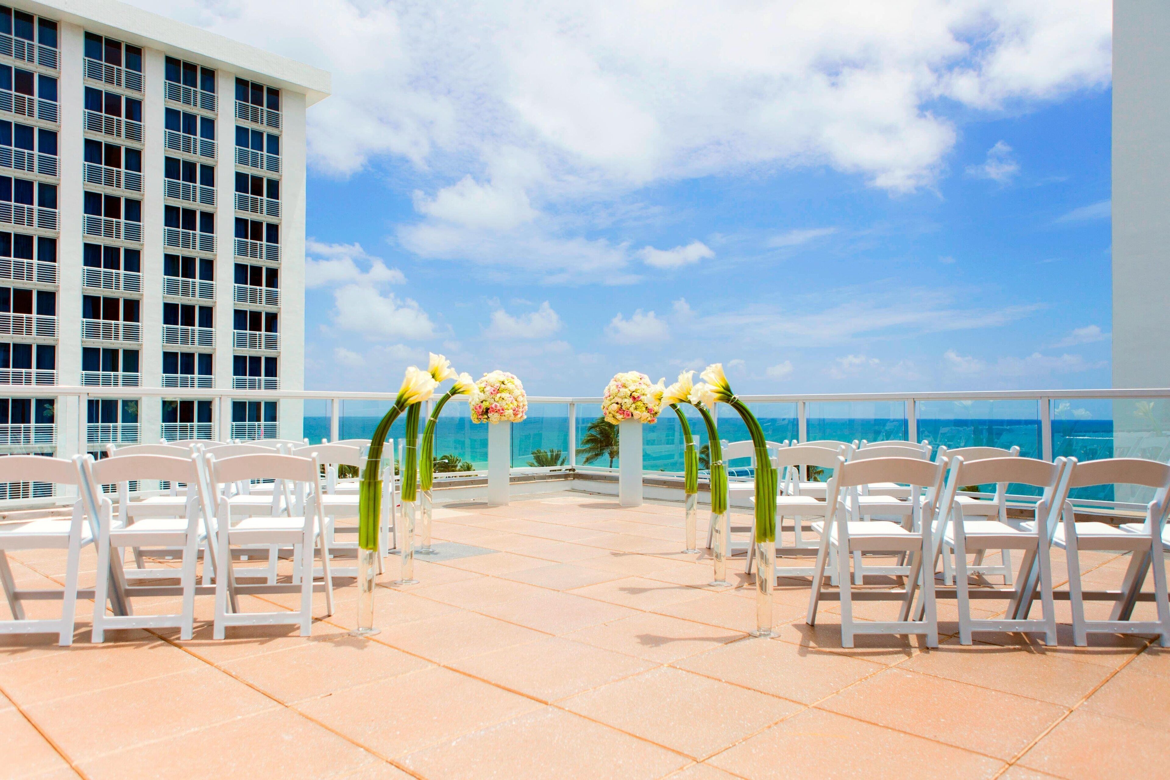 An outdoor ceremony setup with white chairs and floral decorations on a terrace with a view of the ocean and a hotel building in the background.
