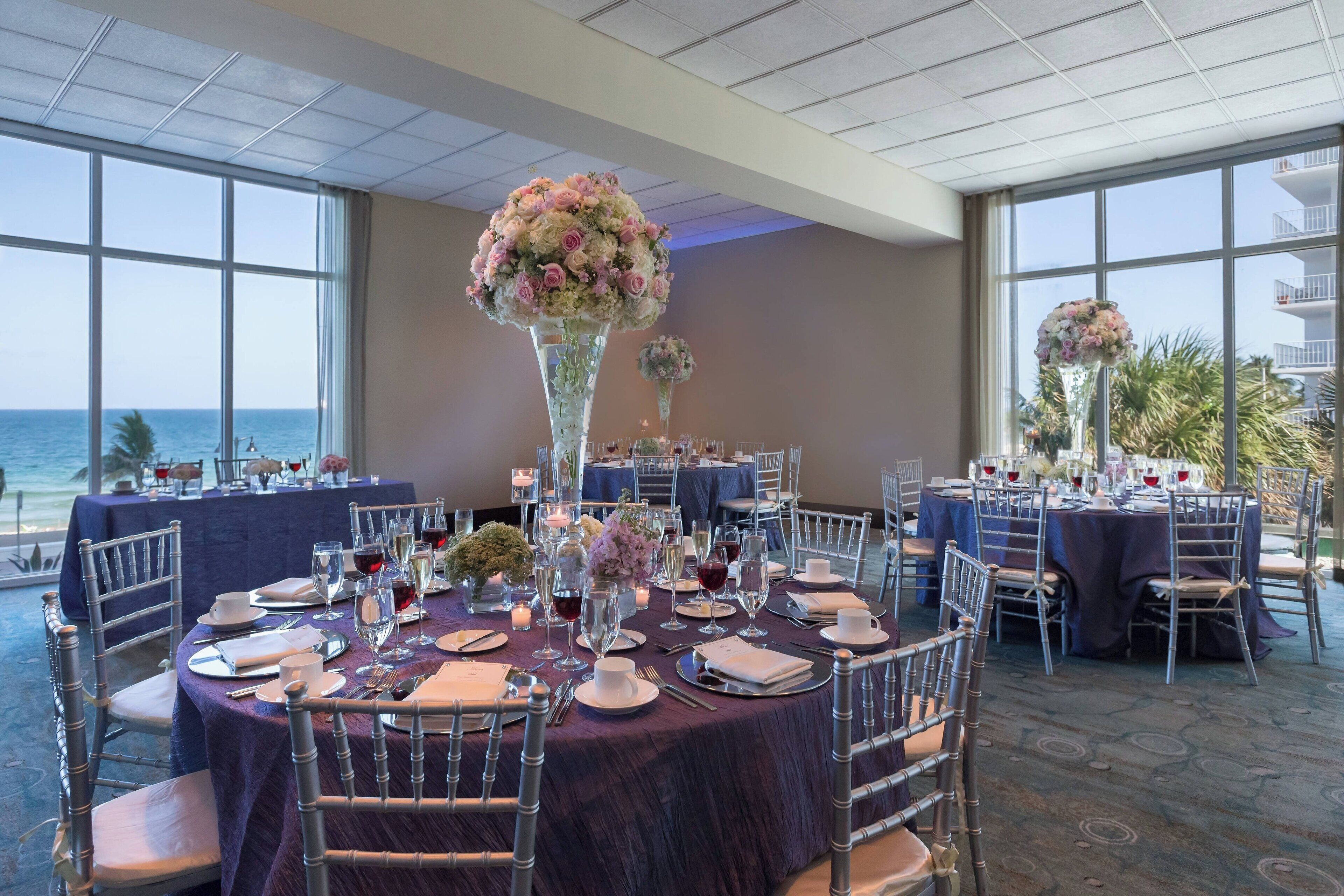 A dining area with round tables set for a formal event, large floral centerpieces, and a view of the ocean through large windows.