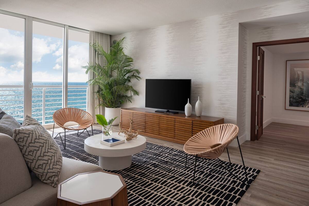 Un salon moderne doté d’une grande fenêtre donnant sur la mer, d’un téléviseur à écran plat posé sur un meuble en bois et de deux fauteuils en osier.