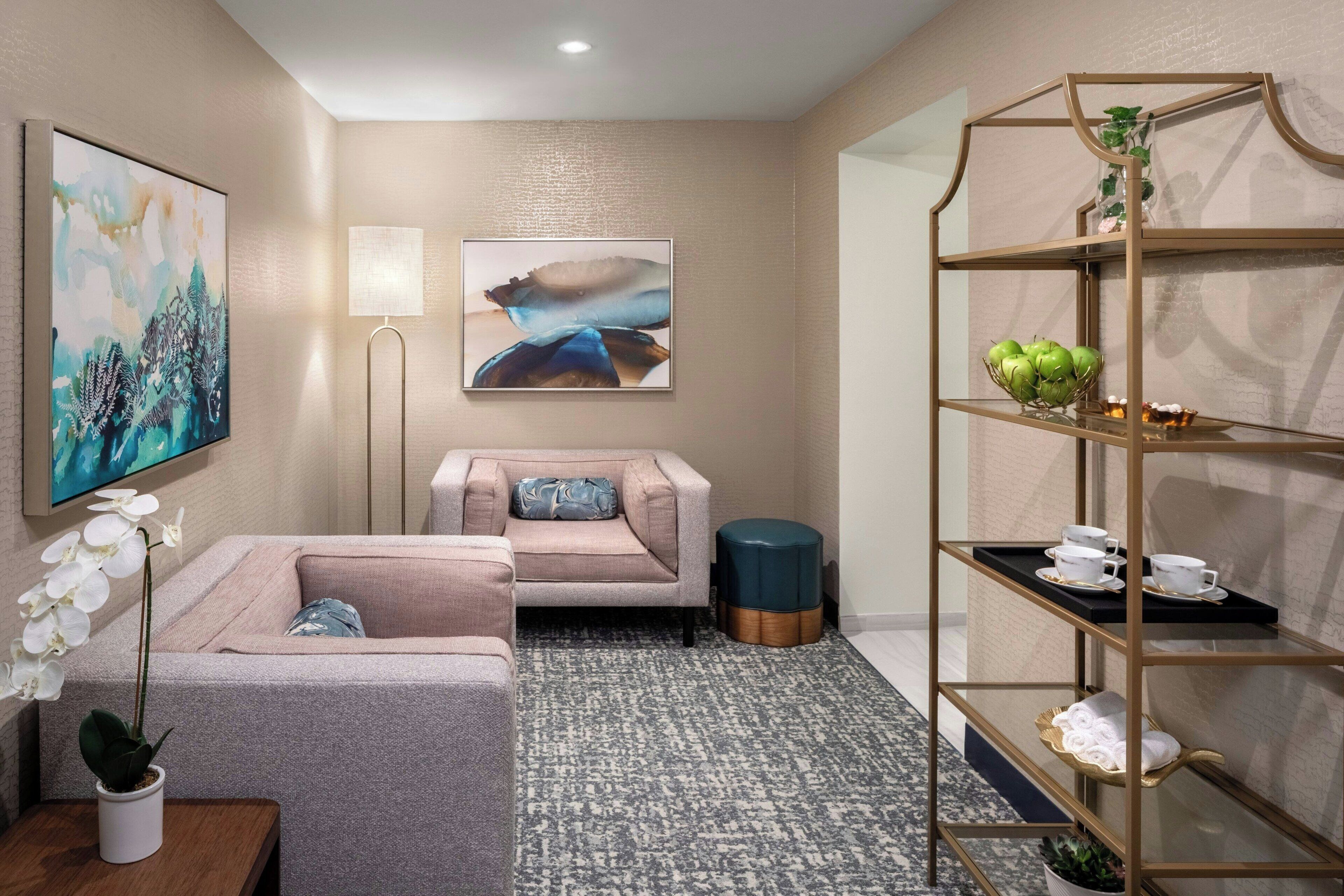 Ein modernes Hotelzimmer mit einem grauen Sofa, einem kleinen runden Tisch mit einem Orchideenstrauß, einer Stehlampe und einem goldenen Metallregal mit Teeservice.