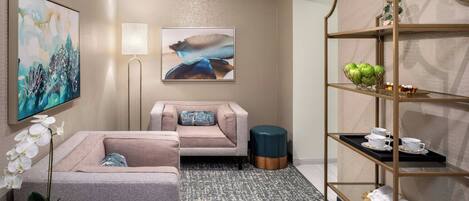 Una habitación de hotel moderna con un sofá gris, una mesita redonda pequeña con una orquídea en maceta, una lámpara de pie y un estante de metal dorado con vajillas de té.
