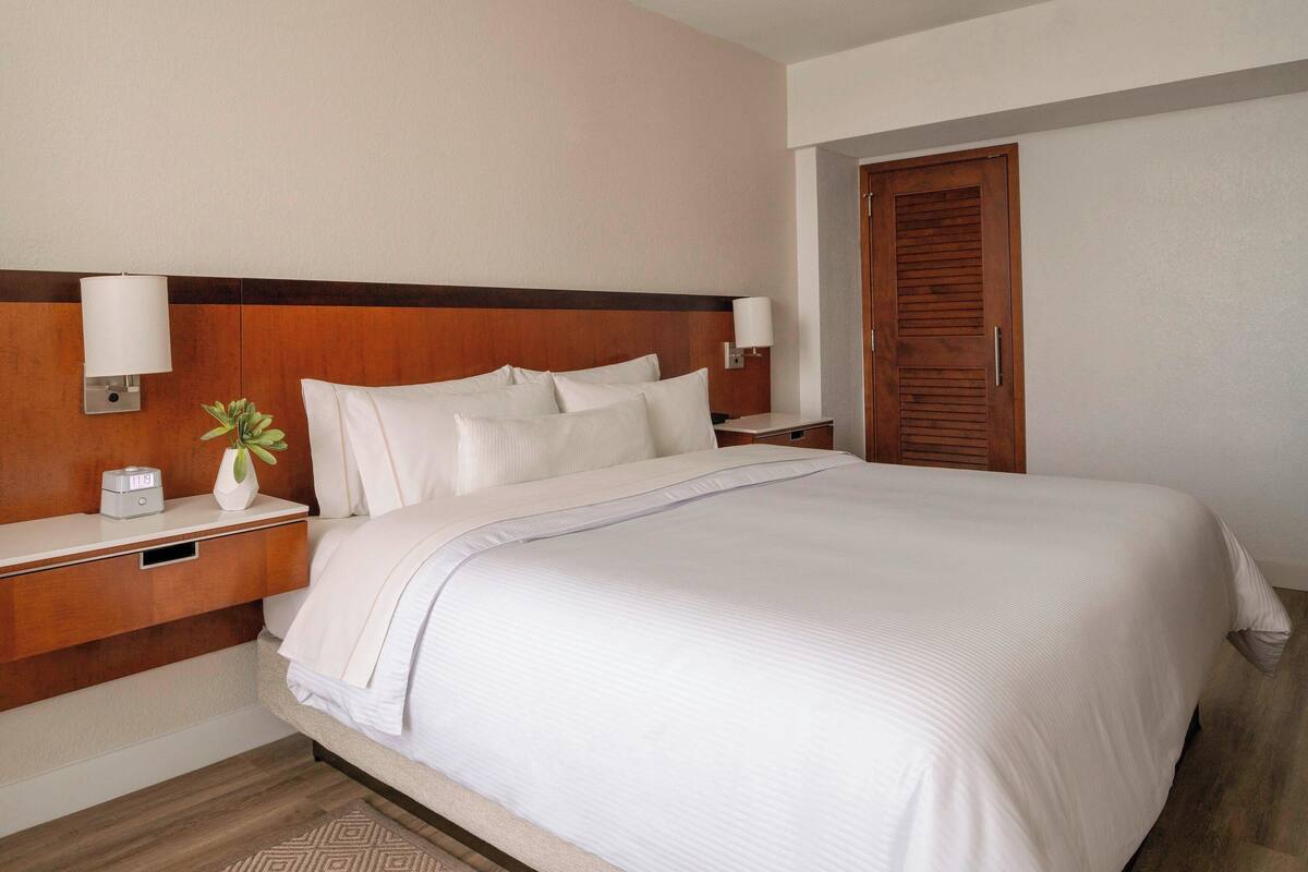 Une chambre d’hôtel avec un grand lit, deux tables de chevet, une petite table, une plante en pot et une porte en bois.