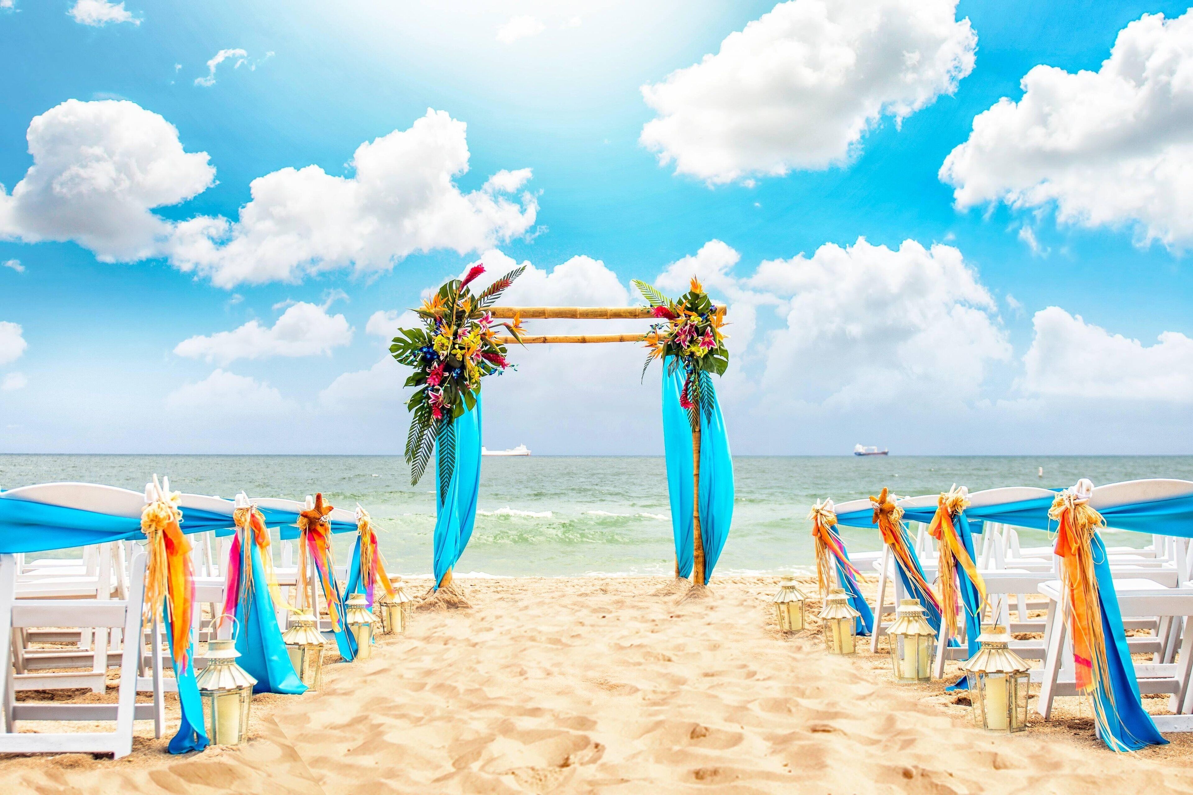 A beach wedding setup with a wooden arch decorated with flowers and blue drapes, white chairs lined up on the sand, and a view of the ocean.