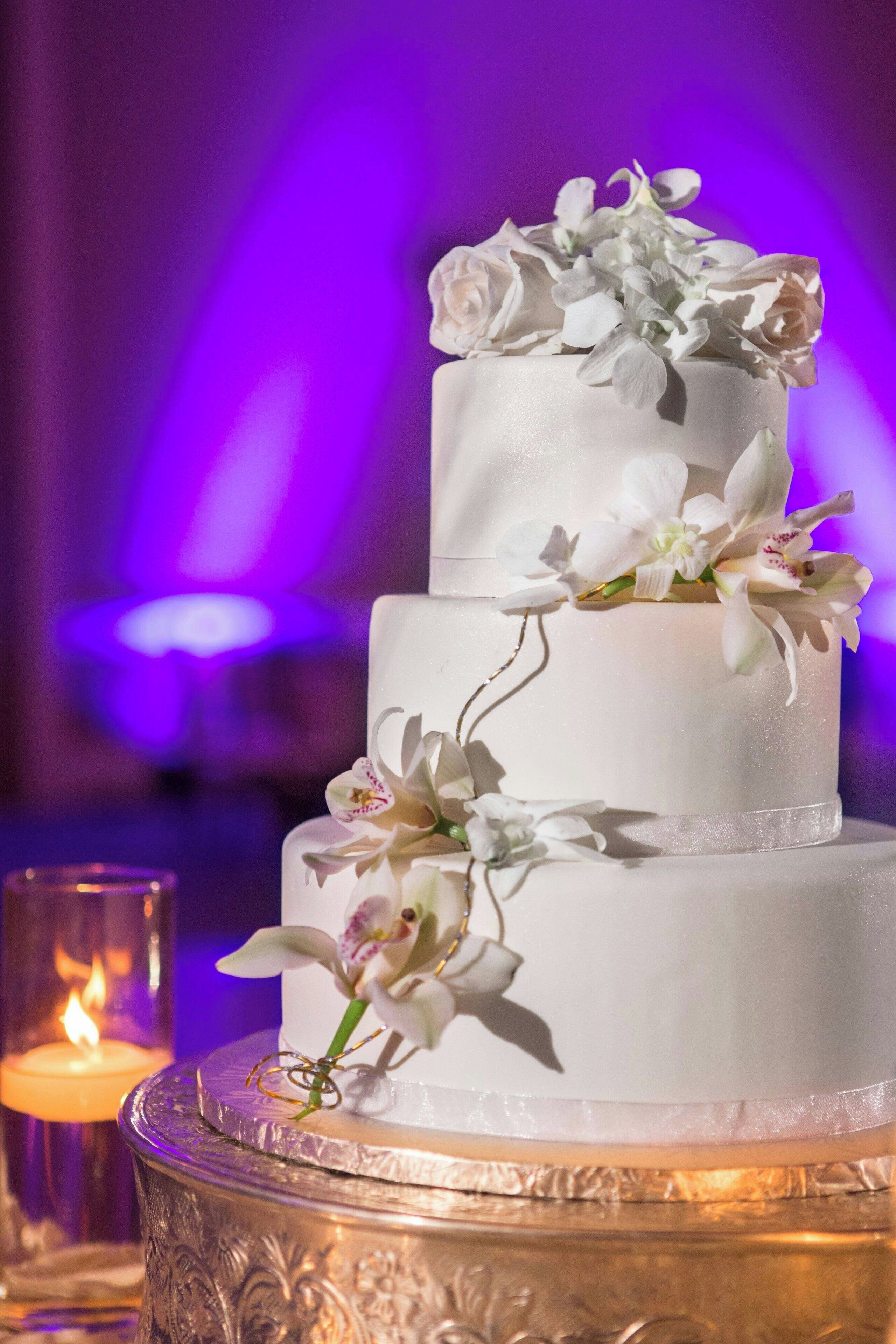 Un pastel blanco de varios niveles decorado con flores, sobre una mesa de vidrio con diseños intrincados, en un ambiente tenuemente iluminado con luces púrpuras.