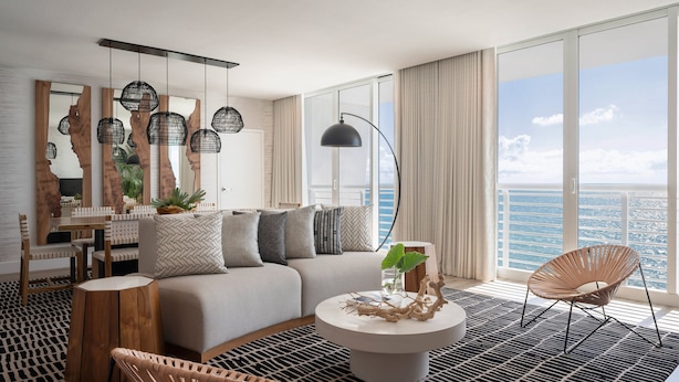 A modern living room with a sofa, coffee table, and chairs, a dining area with a white fence, and large windows with a view of the ocean.