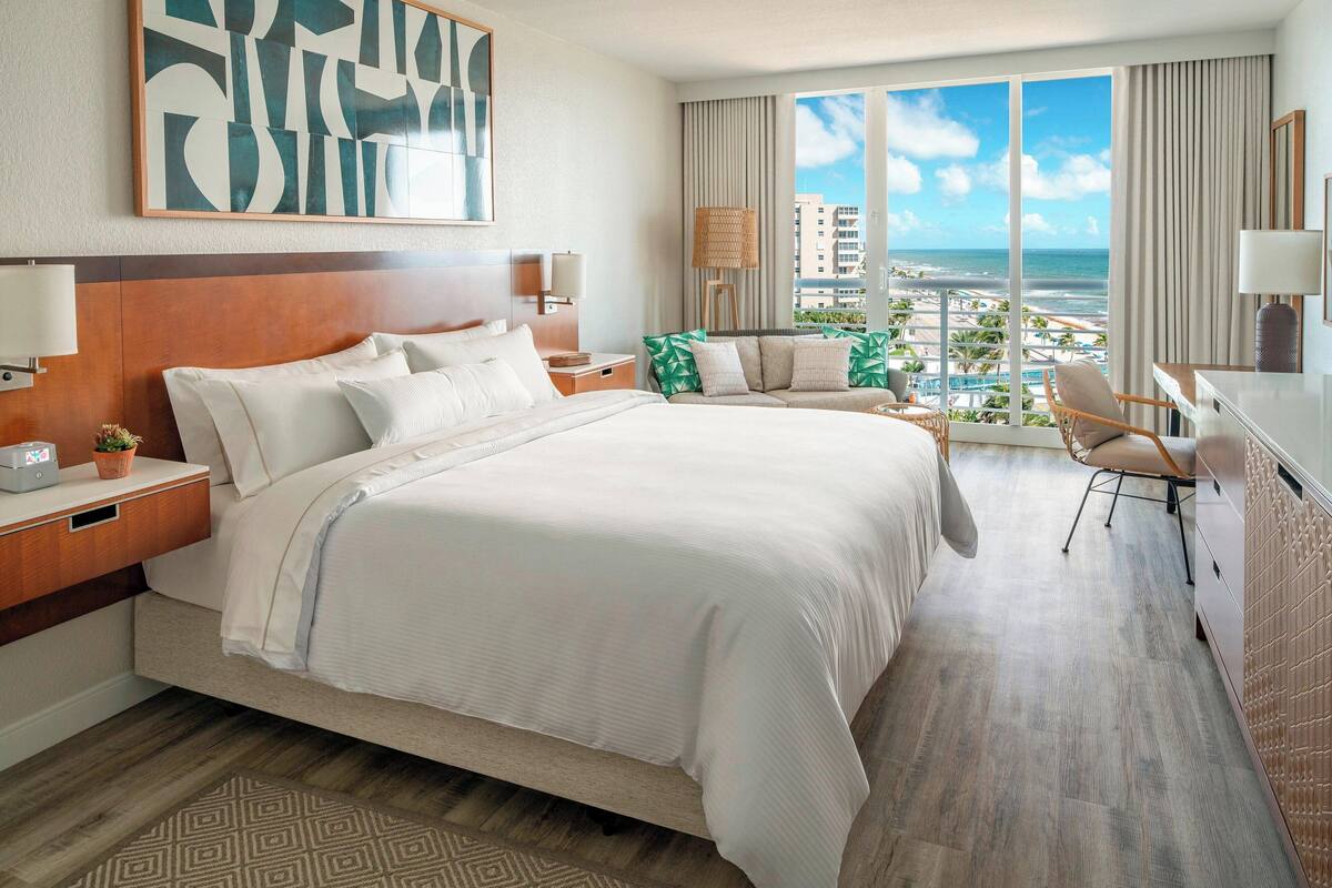 Une chambre d’hôtel avec un grand lit, un bureau, une chaise, une vue sur l’océan et un panorama sur la ville.
