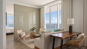 A modern hotel room with a large window offering a sea view, a sofa, a dining table, and wicker chairs.