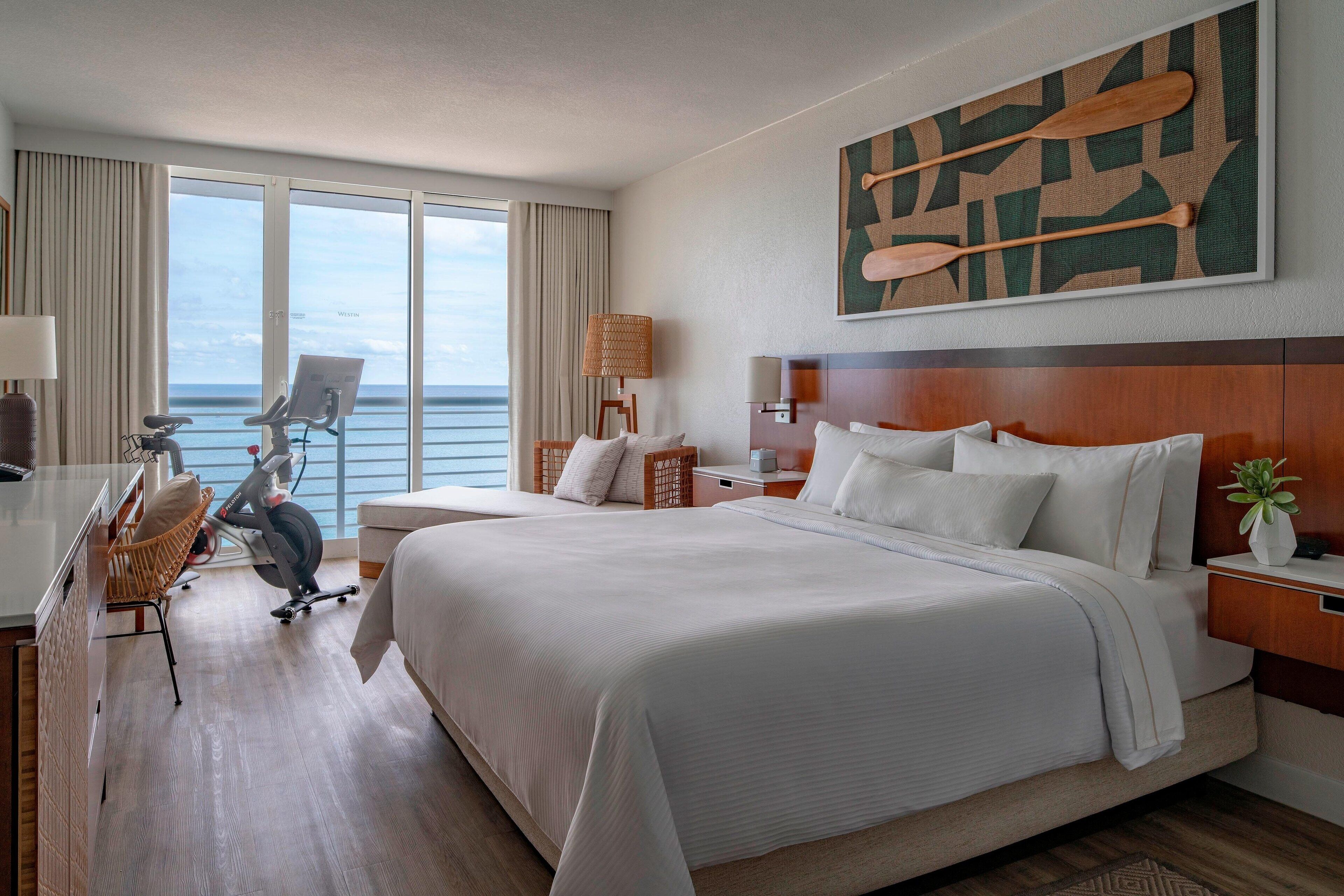 A hotel room with a large bed, a desk with a chair, a treadmill, and a view of the ocean.