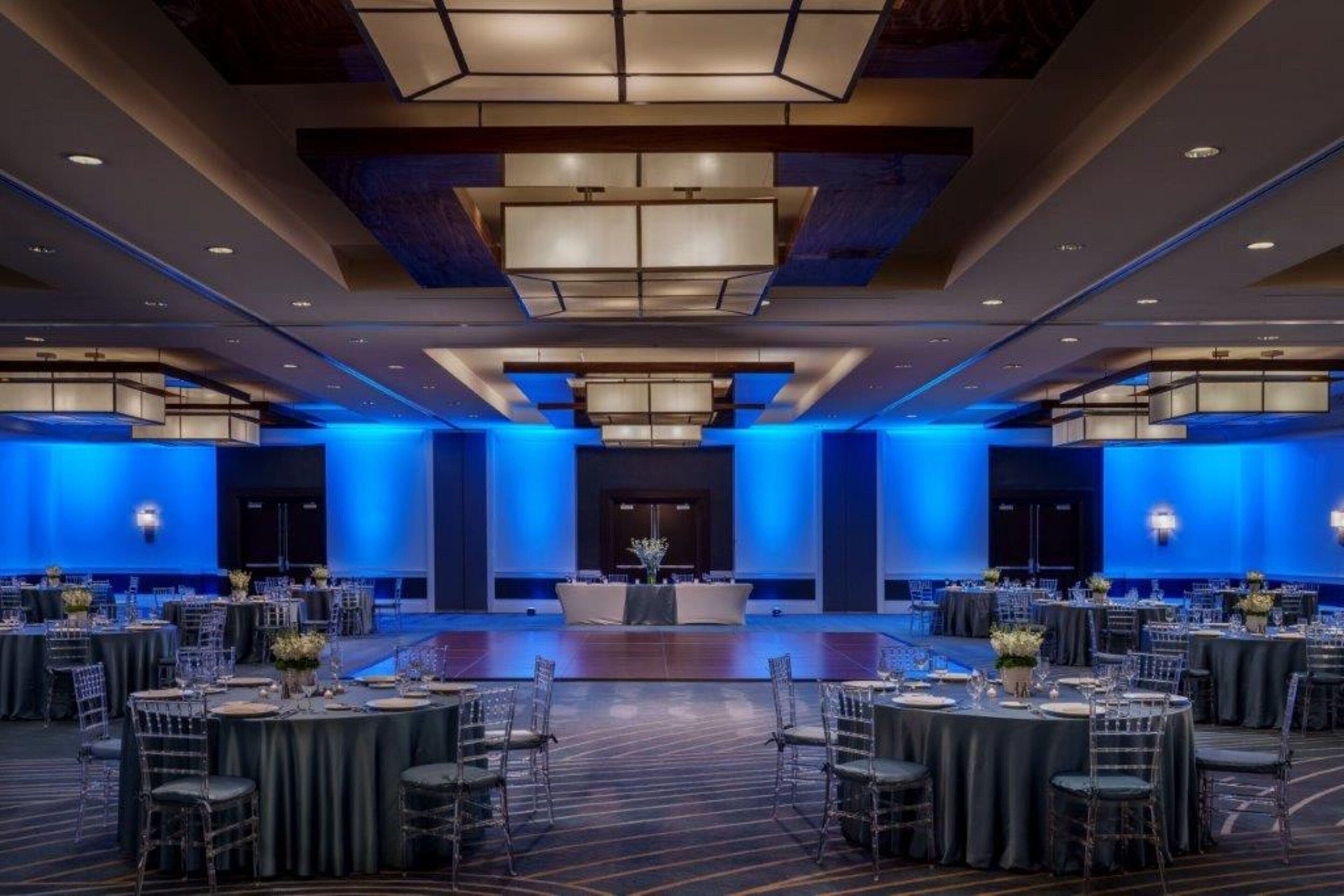 Un amplio salón de banquetes con mesas y sillas redondas, iluminación ambiental azul y un escenario central.