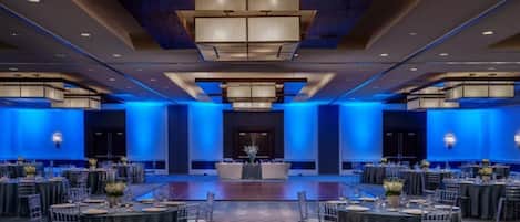 Un amplio salón de banquetes con mesas y sillas redondas, iluminación ambiental azul y un escenario central.
