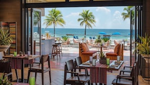 A restaurant with outdoor seating overlooking a beach, featuring palm trees and a view of the ocean.