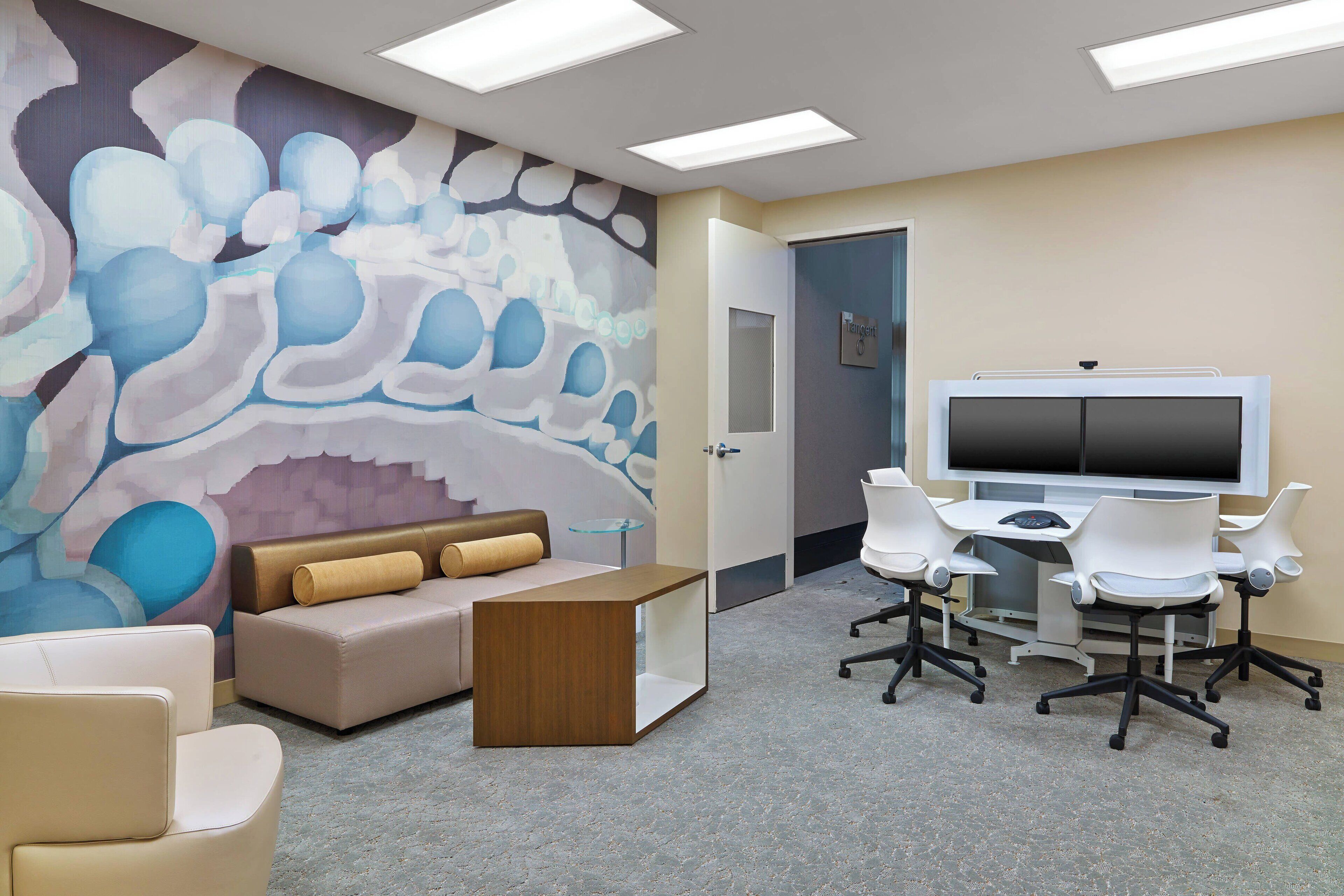 A modern office space with a large abstract mural, a comfortable sofa, a wooden side table, a desk with chairs, and a large flat-screen TV.