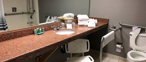 A hotel bathroom with a granite countertop sink, a mirror, a white chair, a toilet, and a shower area.