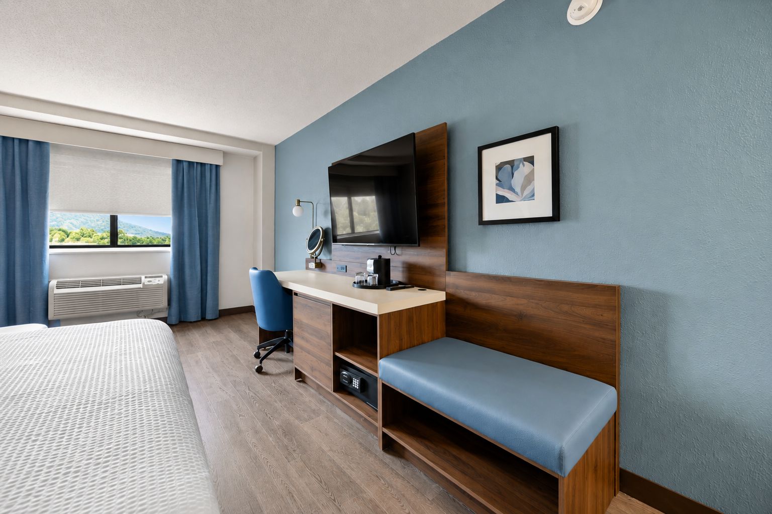 Quarto de hotel com cama, escrivaninha com computador, cadeira, televisão e janela com cortinas.