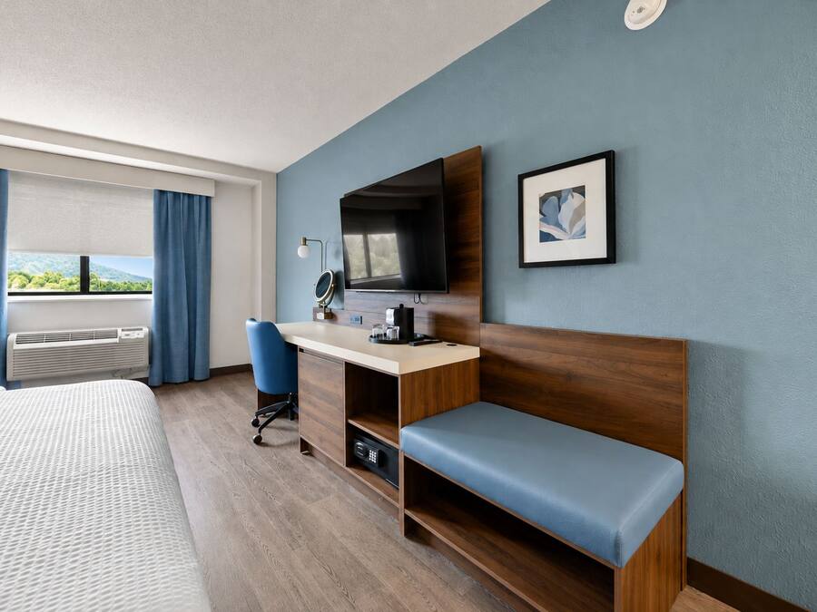 A hotel room with a bed, a desk with a computer, a chair, a television, and a window with curtains.