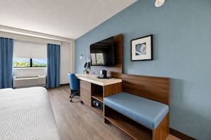 Una habitació d'hotel amb un llit, un escriptori amb un ordinador, una cadira, una televisió i una finestra amb cortines.