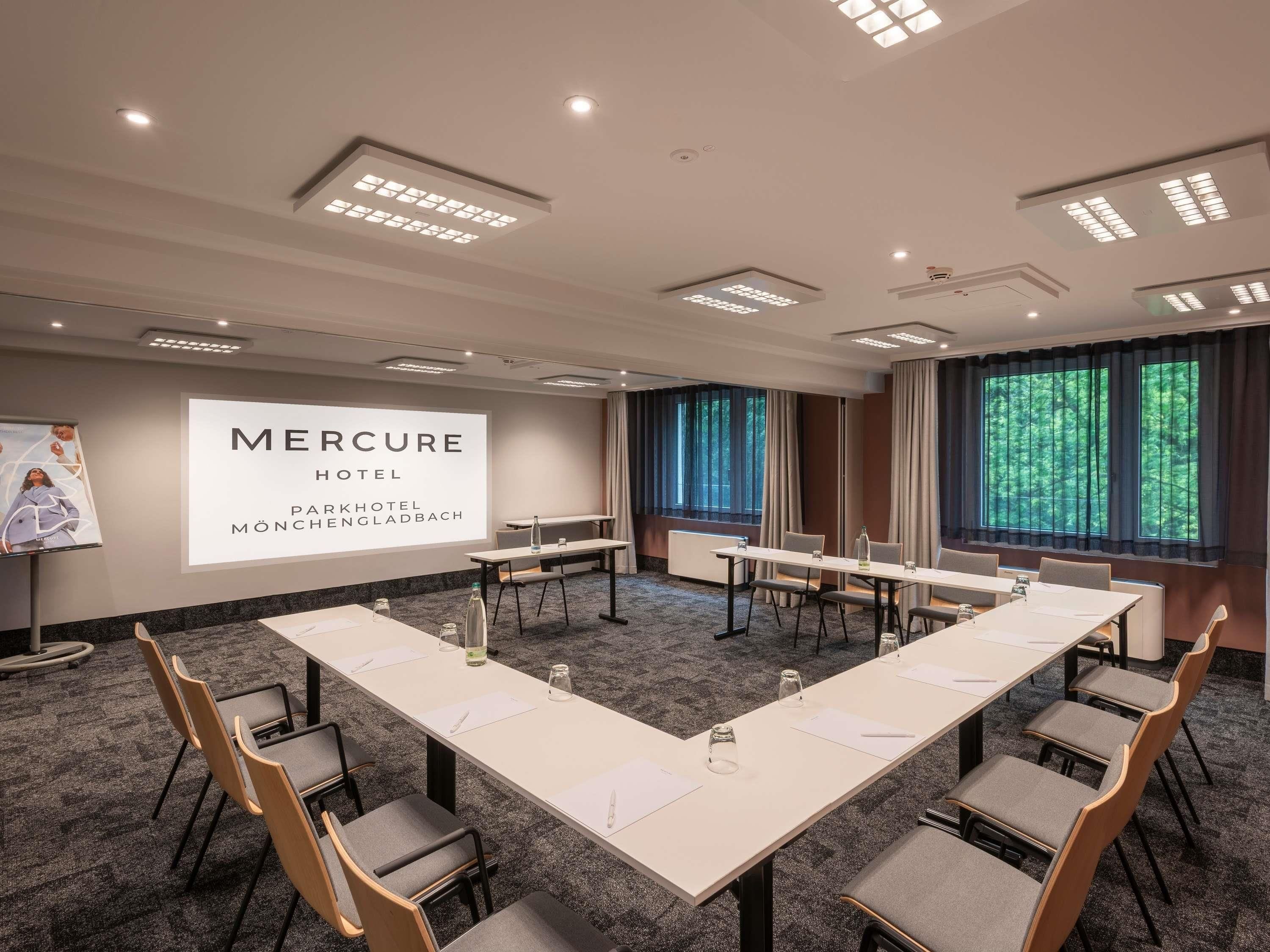 A conference room with a long table, chairs, a projector screen, and a view of trees through the windows.