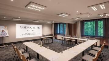 A conference room with a long table, chairs, a projector screen, and a view of trees through the windows.
