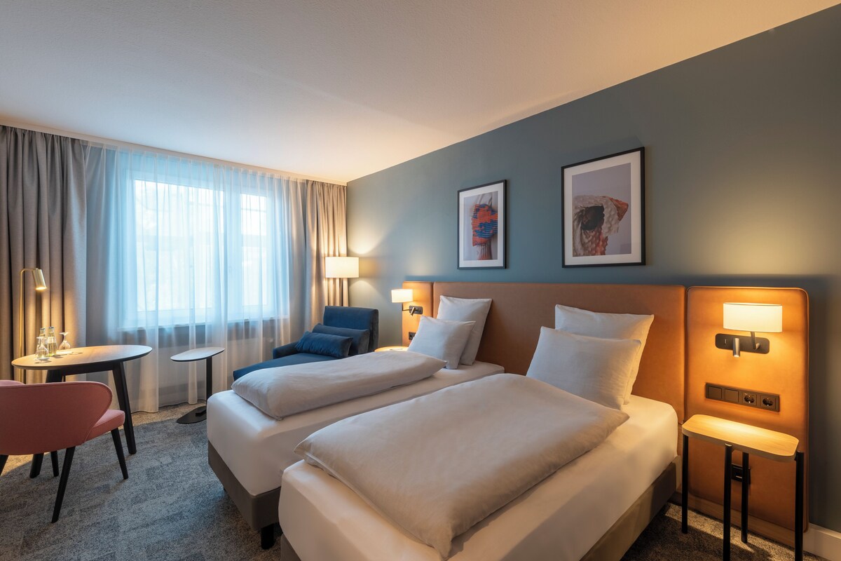 A hotel room with two beds, a desk, a chair, and two wall-mounted lamps.