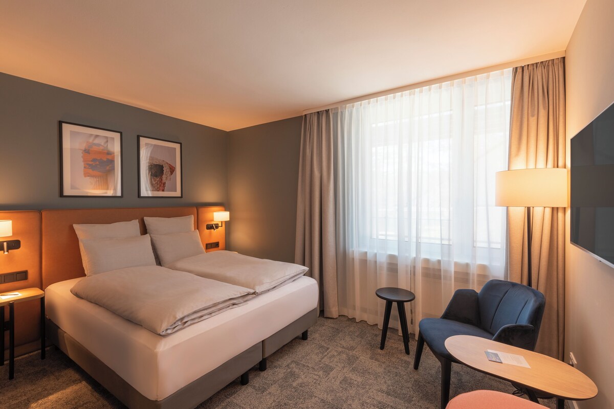 A hotel room with a double bed, two bedside lamps, a small table, a chair, and a large window with curtains.