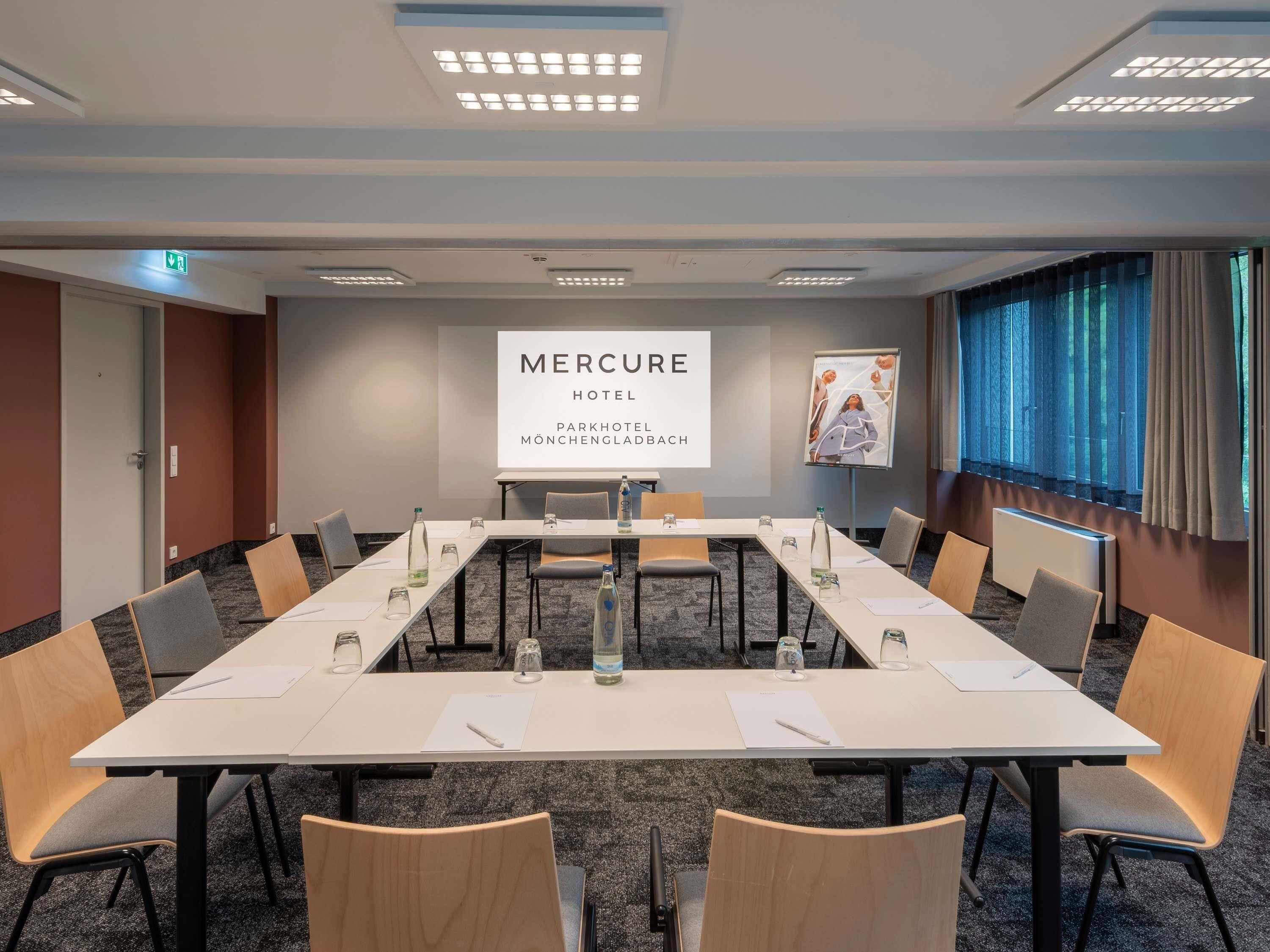 A conference room with a long table, chairs, a projector screen displaying 'MERCURE HOTEL PARKHOTEL MÖNCHENGRAD', and a poster on the wall.