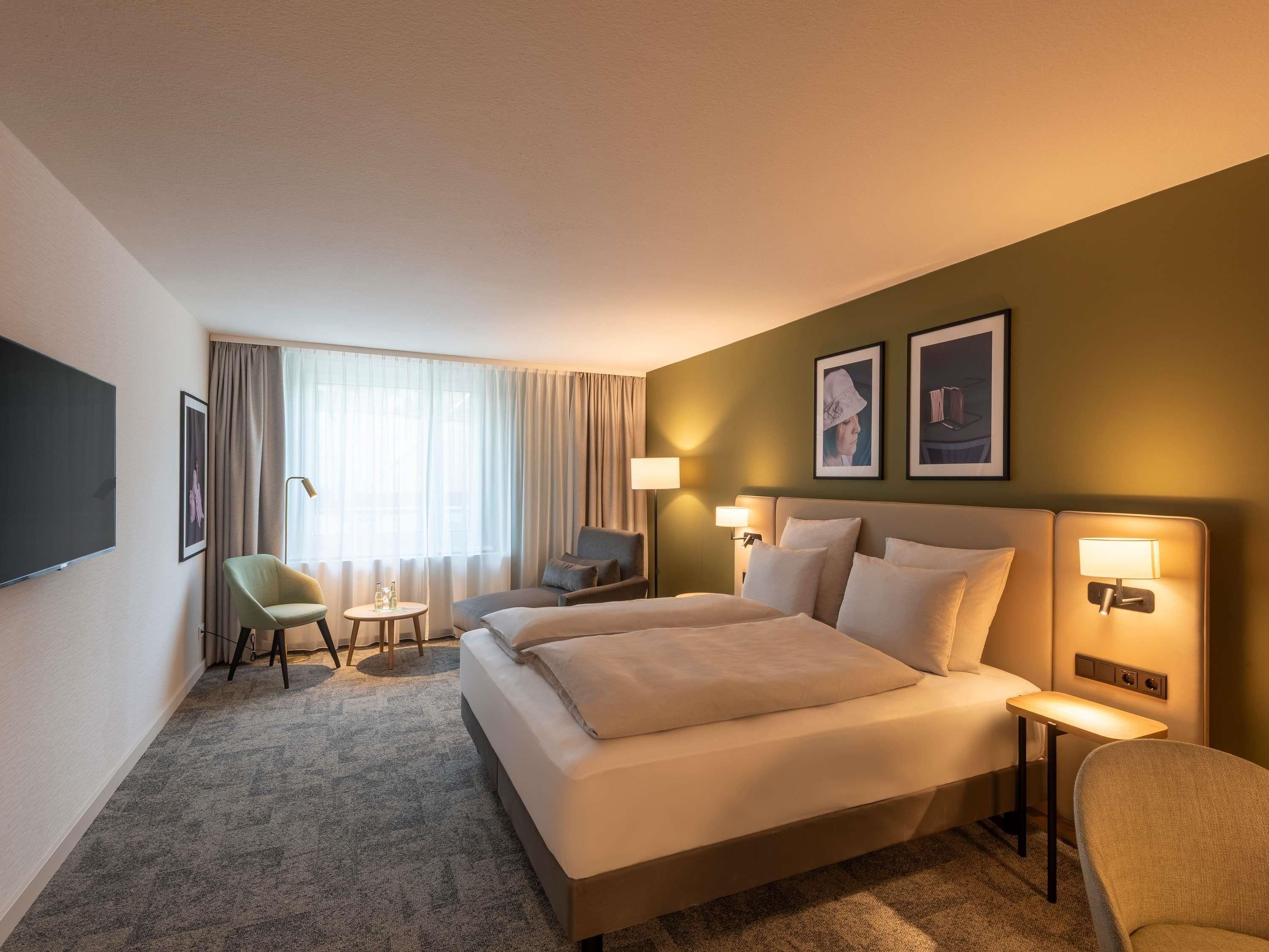 Habitación de hotel con cama, dos mesitas de noche, una silla, escritorio, televisor de pantalla plana y ventana con cortinas.
