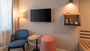 A hotel room with a flat-screen TV, a small wooden side table, a blue armchair, a pink ottoman, and a wall-mounted shelf with a kettle and cups. - Mercure Parkhotel Mönchengladbach (Moenchengladbach)