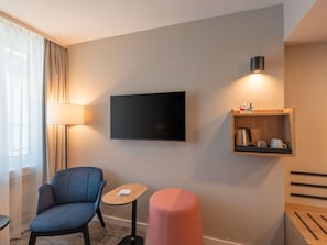 A hotel room with a flat-screen TV, a small wooden side table, a blue armchair, a pink ottoman, and a wall-mounted shelf with a kettle and cups.