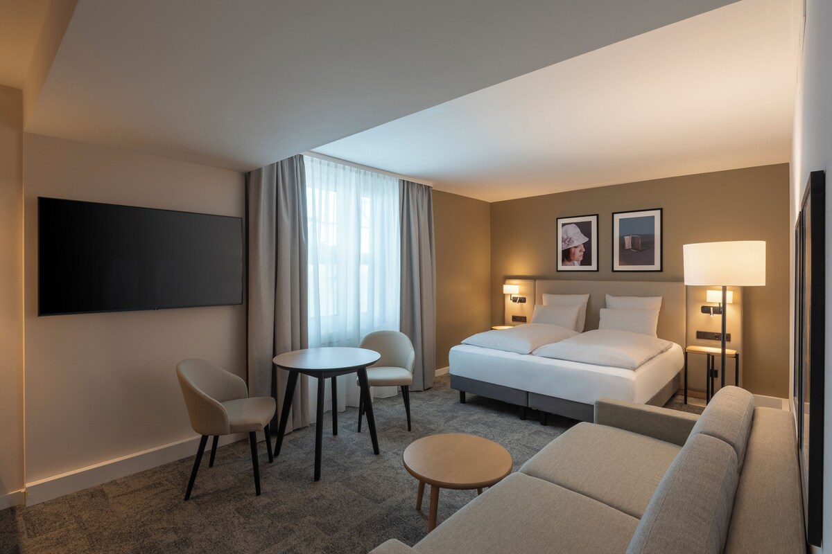 A hotel room with a bed, two chairs, a small table, a sofa, a TV, and two wall-mounted lamps.