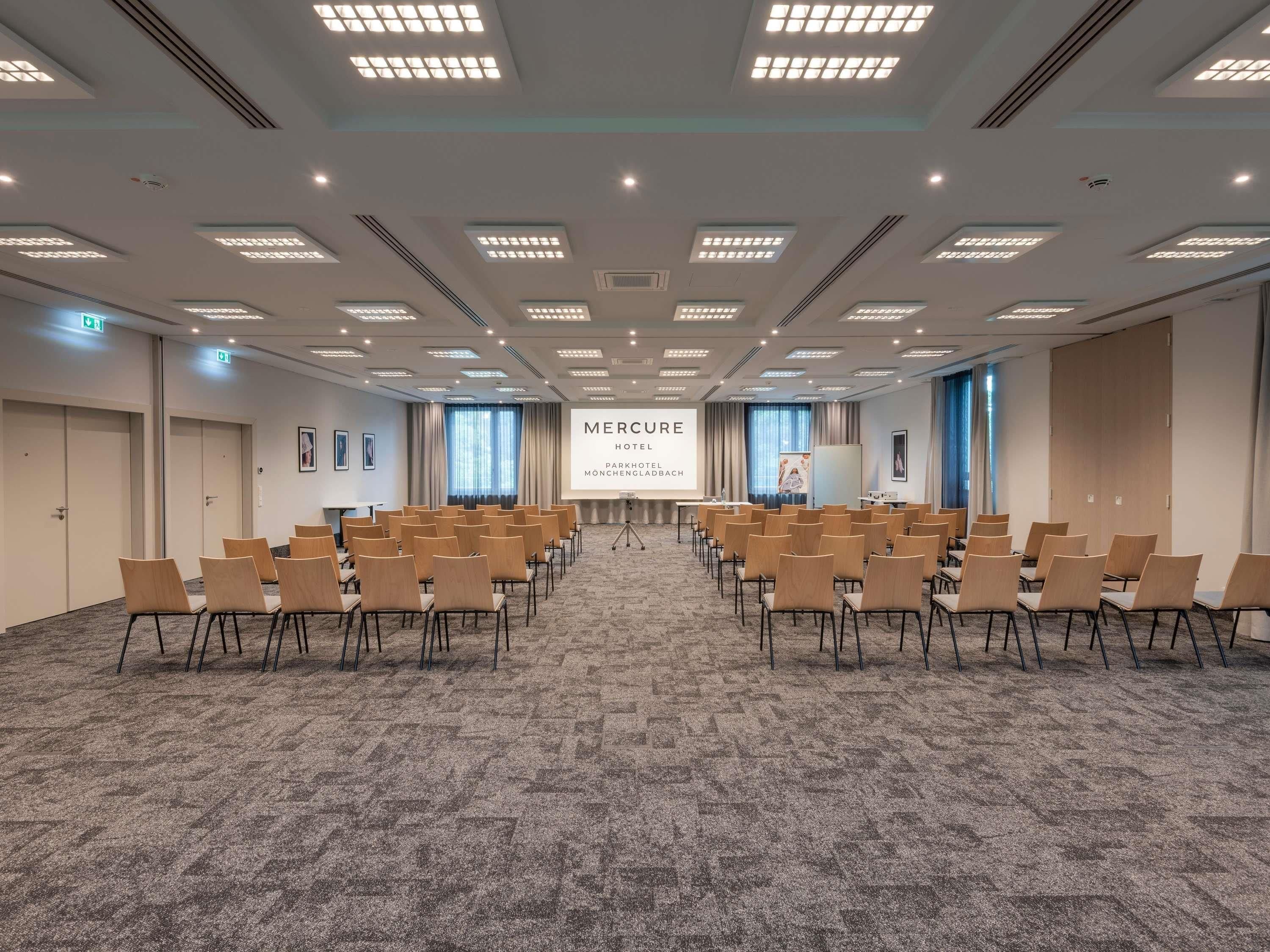 Un salón de conferencias con filas de sillas, una pantalla de proyección y suelo alfombrado.
