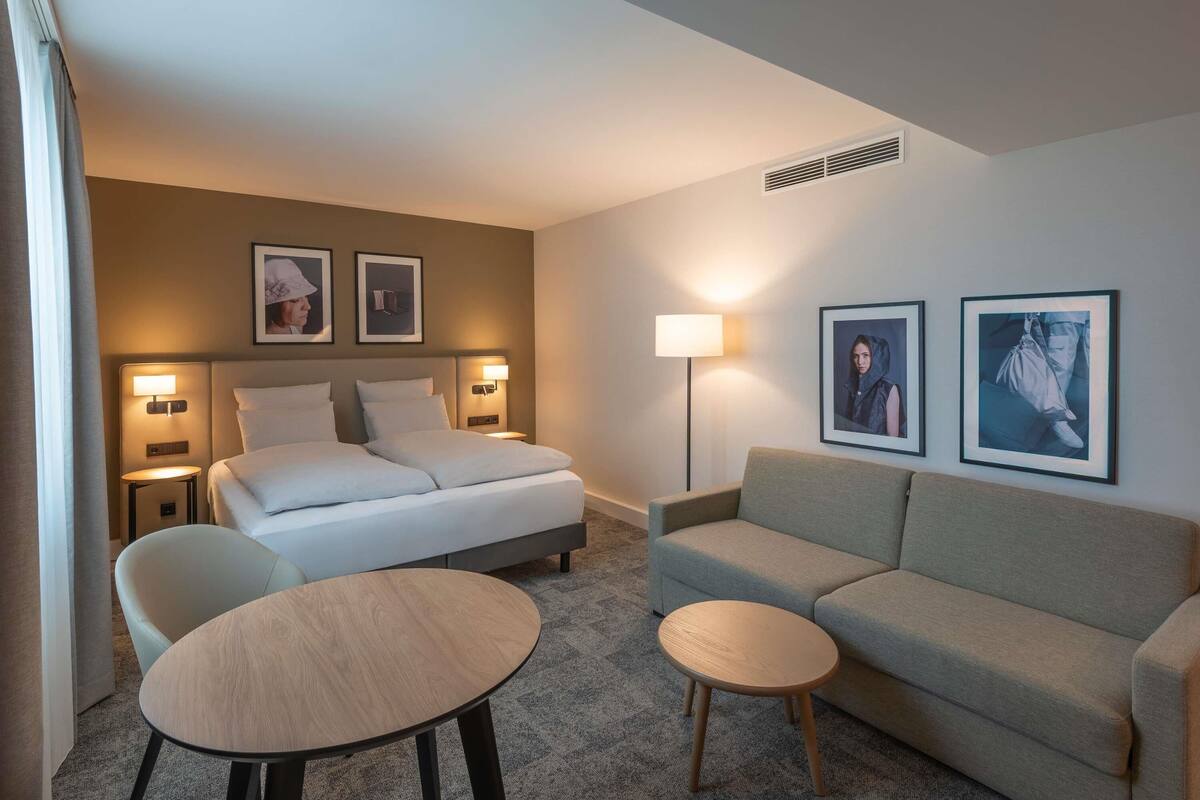 A hotel room with a bed, two bedside tables, a sofa, a small round table, and two wall-mounted lamps.