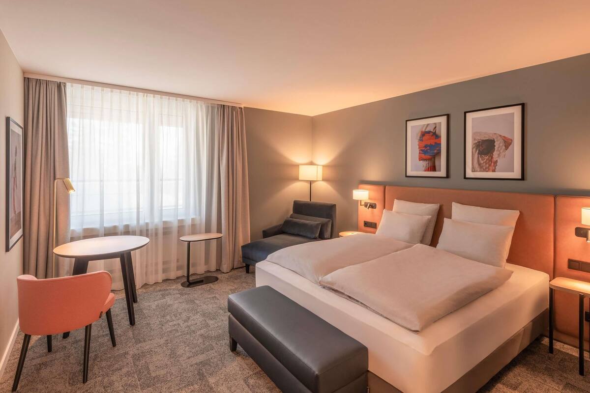 A hotel room with a bed, two bedside tables, a chair, a small table, and a window with curtains.