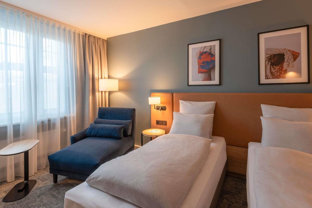A hotel room with a bed, a sofa, a small table, a lamp, and two framed pictures on the wall.