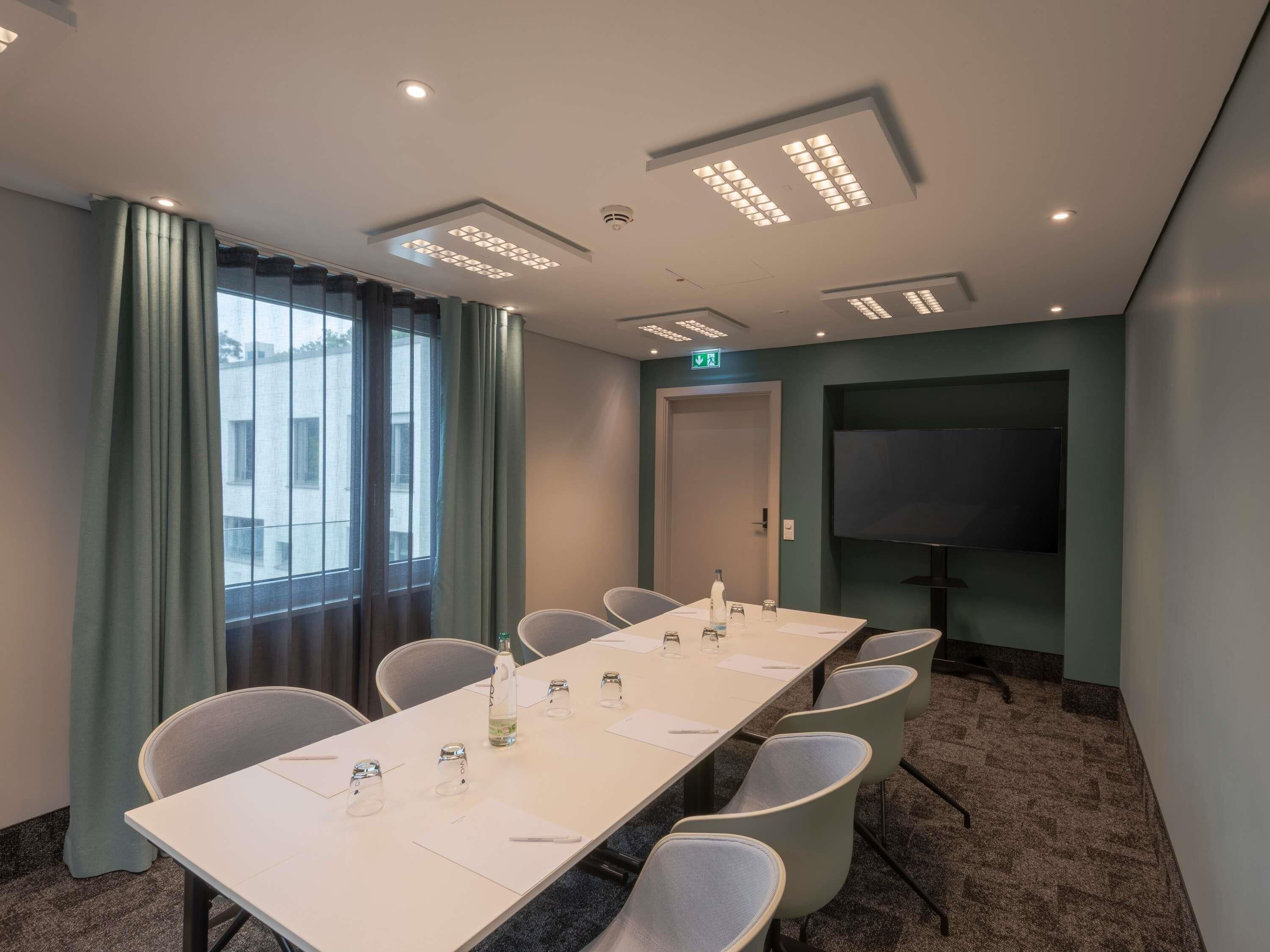 Une salle de conférence avec une longue table, des chaises, un écran de projection et une porte avec un panneau indiquant la sortie.