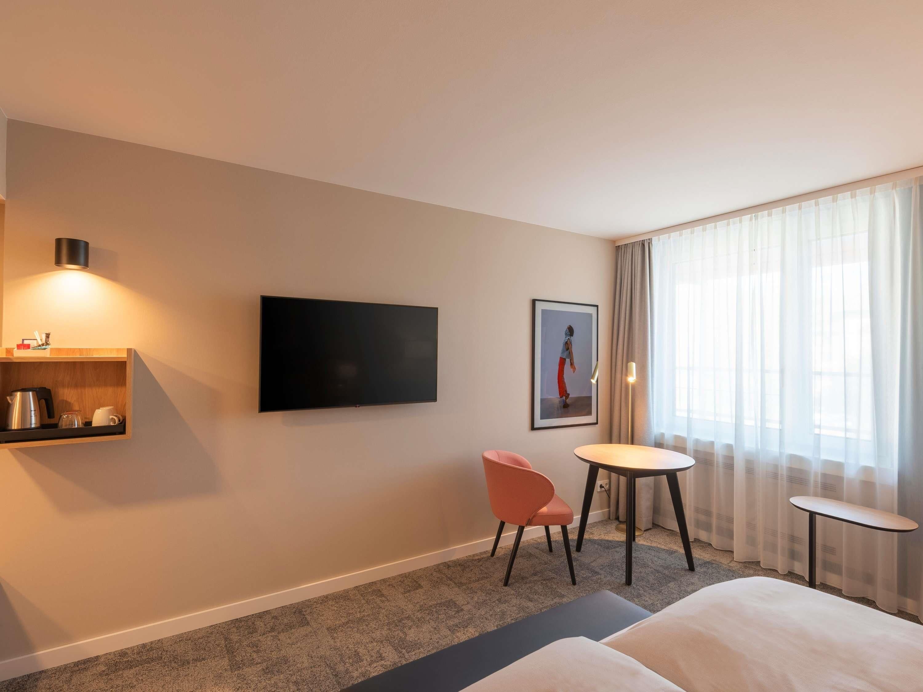 A hotel room with a flat-screen TV, a small table, a chair, a sofa, and a picture on the wall.