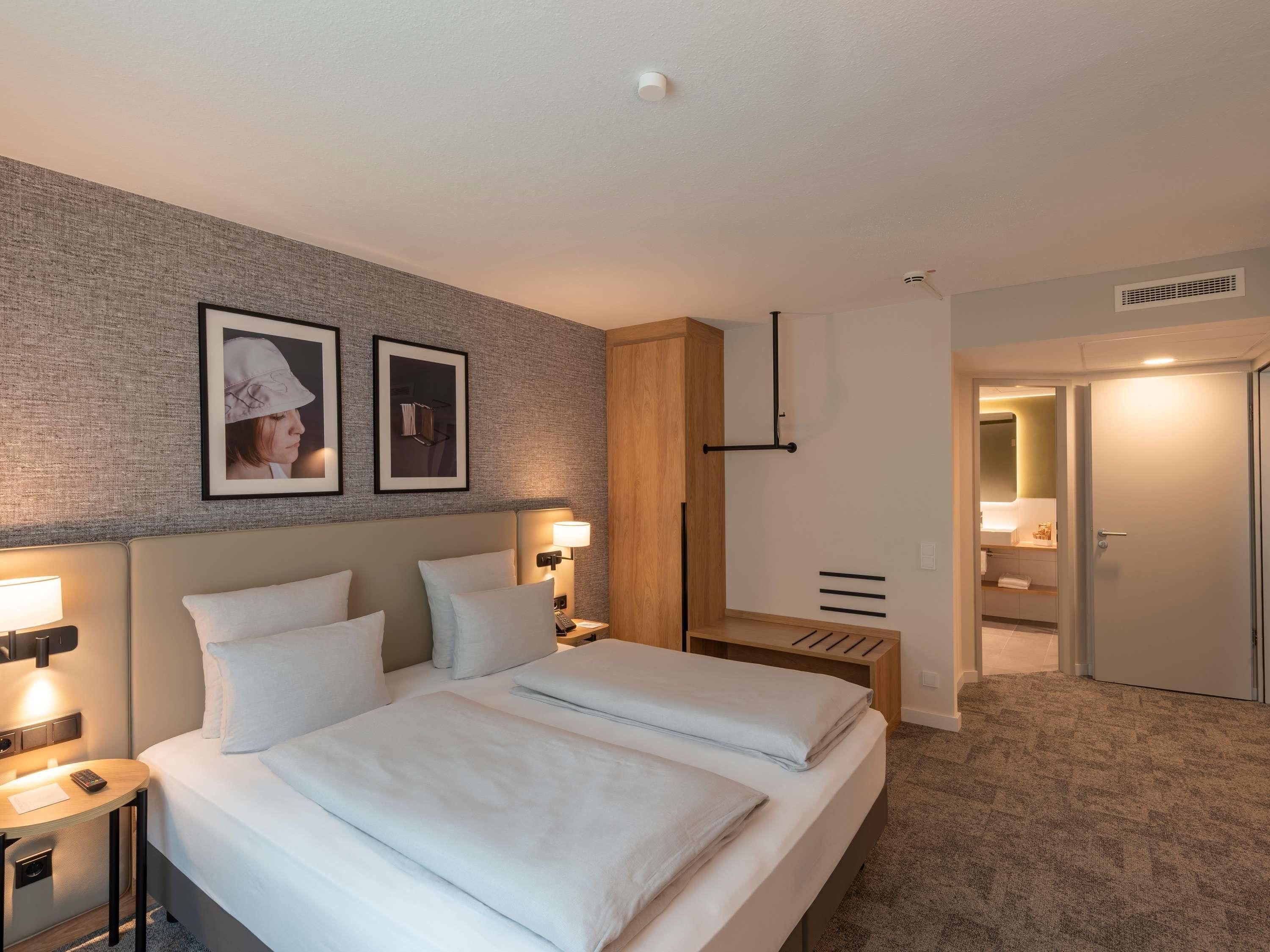 Habitación de hotel con cama doble, mesitas de noche, lámpara, cuadro enmarco, mueble de madera, espejo y puerta que da al baño.