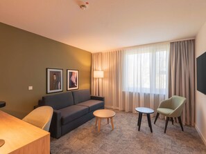 A hotel room with a sofa, two chairs, a small round table, a lamp, and two framed pictures on the wall.