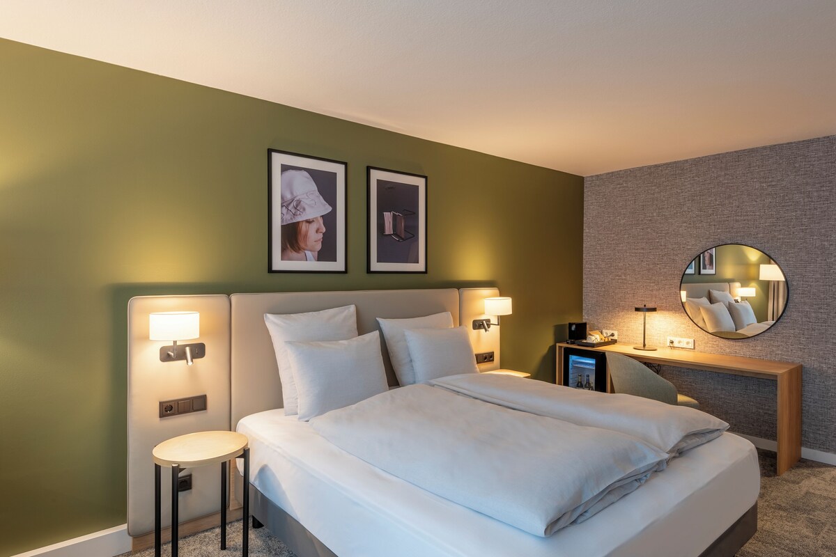 A hotel room with a bed, bedside lamps, a small table, a desk with a lamp, and two framed pictures on the wall.