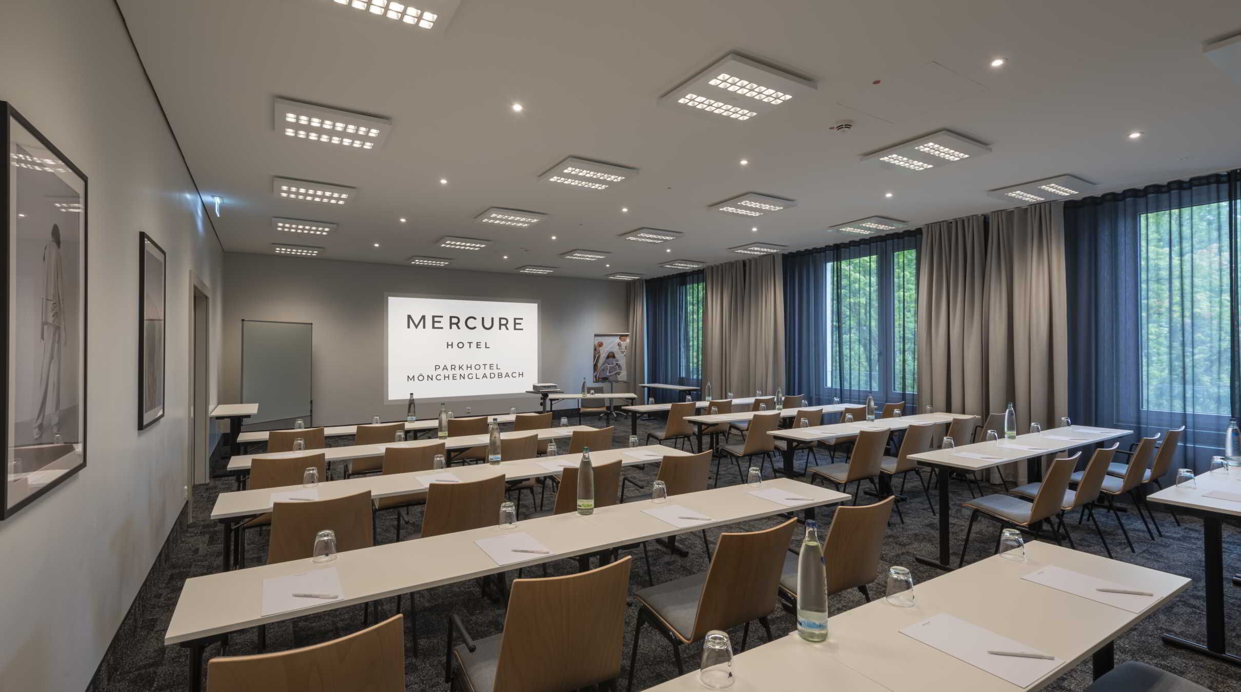 Une salle de conférence équipée de tables, de chaises et d’un écran de projection affichant le texte suivant : « MERCURE HOTEL PARKHOTEL MÖNCHENGLADBACH ».