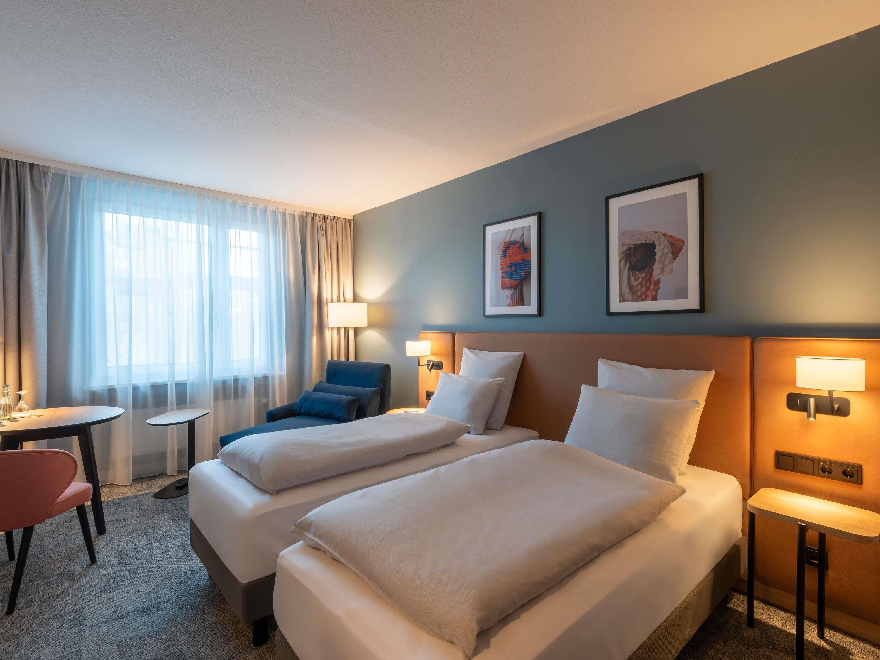A hotel room with two beds, a desk, a chair, and two wall-mounted lamps.