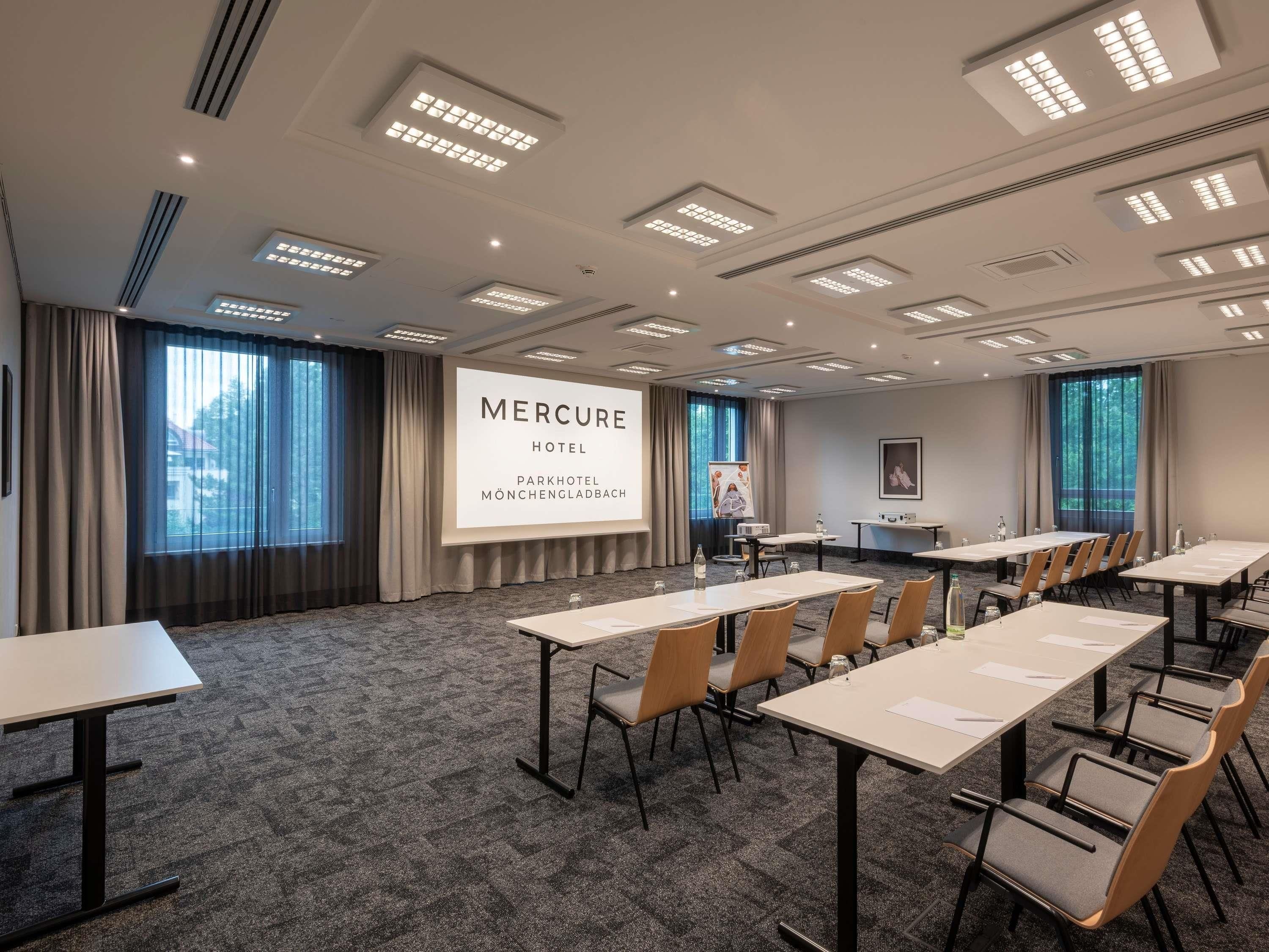 Un salón de conferencias con varias mesas y sillas, una pantalla grande mostrando 'Mercure Hotel Parkhotel Mönchengladbach' y amplias ventanas con cortinas.