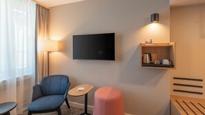 A hotel room with a flat-screen TV mounted on the wall, a small wooden table, a blue armchair, a pink ottoman, and a shelf with a kettle and cups. - Mercure Parkhotel Mönchengladbach (Moenchengladbach)