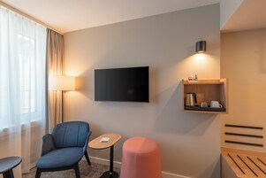 Habitación de hotel con televisor de pantalla plana montado en la pared, una mesita de madera, un sillón azul, un otoman rosa y una repisa con hervidor y tazas.