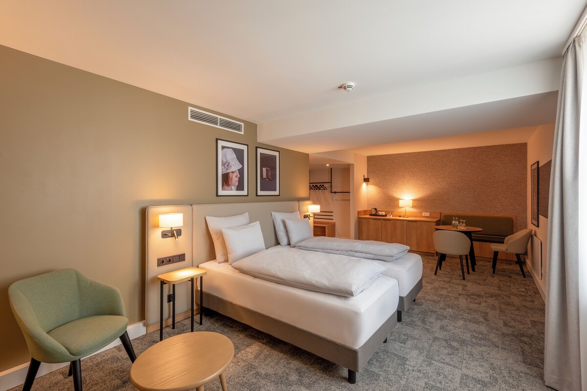 A hotel room with two beds, a desk, a chair, a round table, and a small kitchenette.