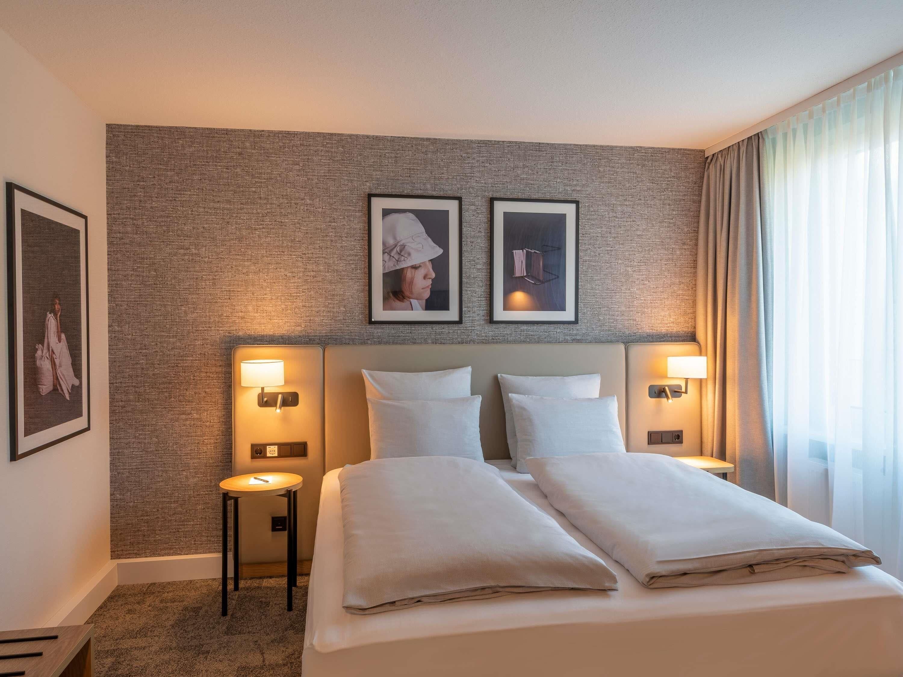 Hotellihuone, jossa on suuri sänky, kaksi yöpöytää, seinään kiinnitetyt lamput ja seinällä olevia tauluja.